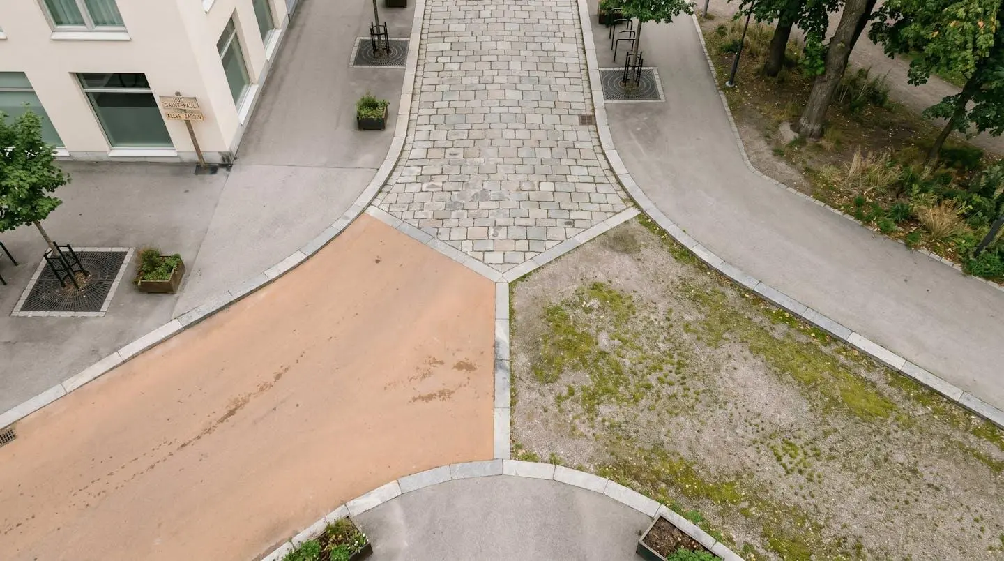 Vue aérienne d'un carrefour urbain où trois types de revêtements de sol se rejoignent : pavés de pierre grise, enrobé lisse ocre et gravier végétalisé, formant un Y symbolisant les choix de matériaux.